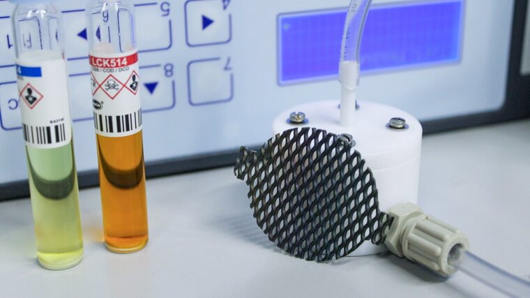 Ein Sensor mit Schlauch-Anschlüssen liegt vor einem elektronischen Gerät und zwei Probenfläschchen mit farbiger Flüssigkeit